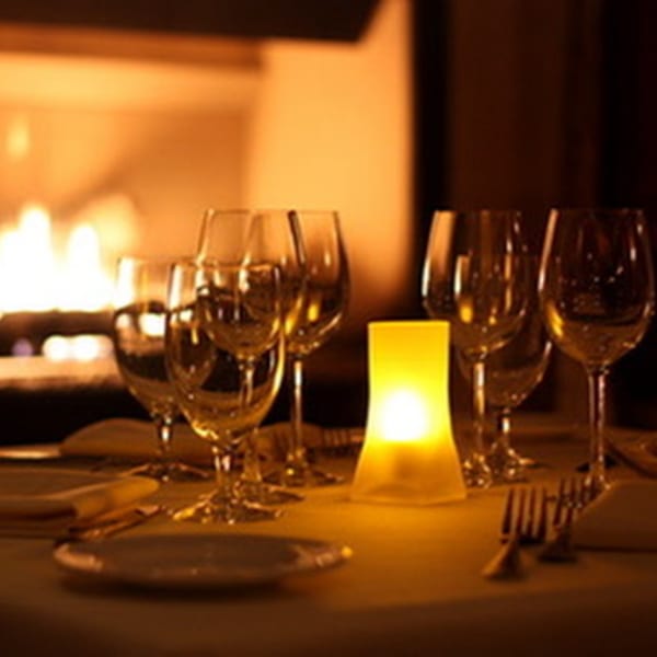 A romantic Luciano's dining table set with wine glasses, a glowing candle, and a fireplace in the background.