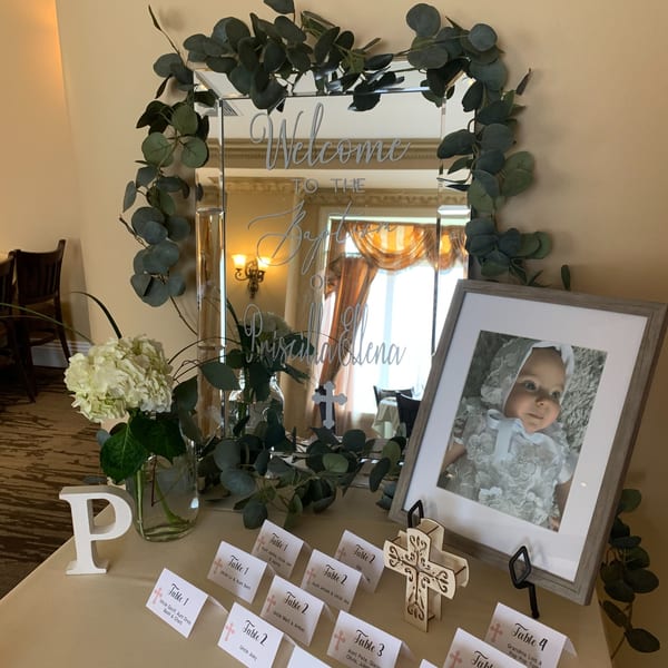 A welcome mirror with eucalyptus, a baby's framed photo, and table setting cards for a baptism.