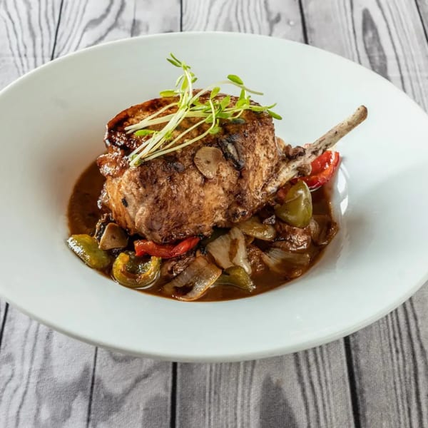 A thick pork chop with a bone, topped with microgreens, served in a rich sauce with peppers and onions.
