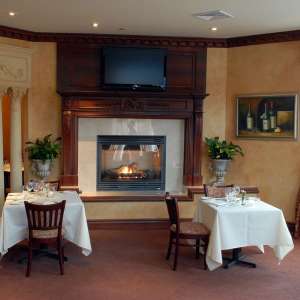 Dining room at Luciano's Ristorante with two set tables, a lit fireplace, and a painting of wine bottles.