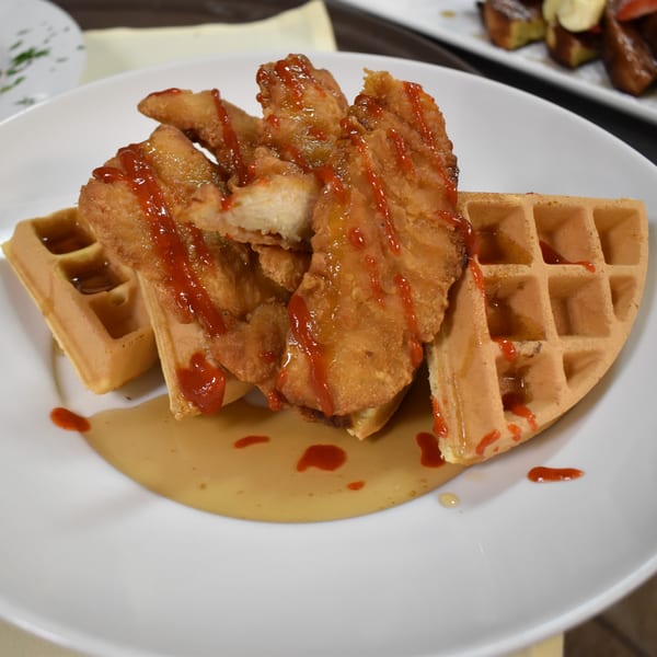 Golden fried chicken tenders with red sauce and syrup on waffles, on a white plate.