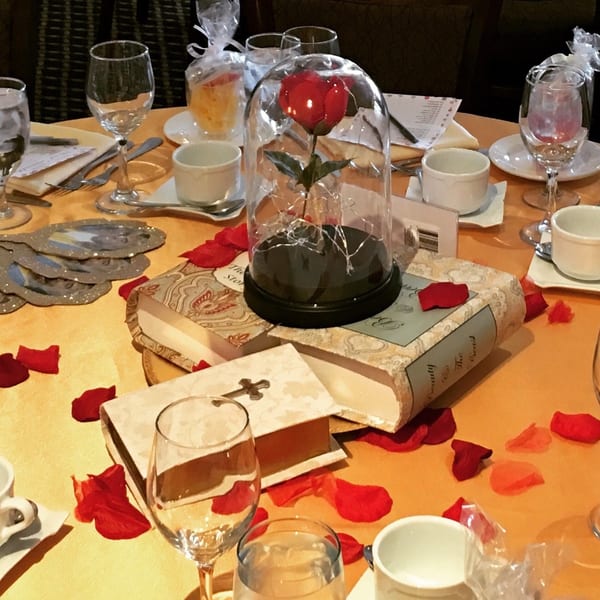 Table set with wine glasses, cups, rose petals, and a Beauty and the Beast enchanted rose centerpiece.