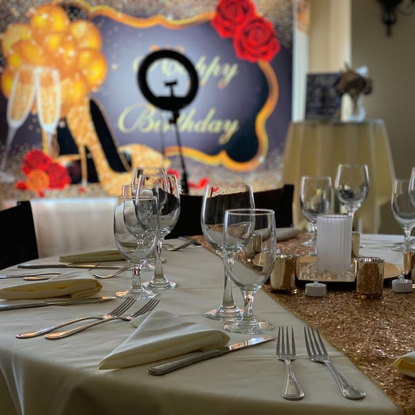 A festive table setting with wine glasses, silverware, and a gold sequin runner, with a "Happy Birthday" backdrop.