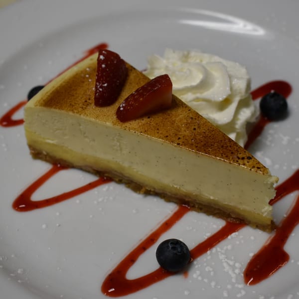 Slice of cheesecake with strawberries, blueberries, whipped cream, and raspberry drizzle on a white plate.