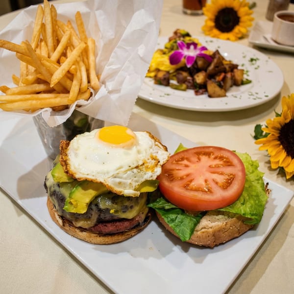 A burger with avocado, melted cheese, and a fried egg, served with fries and a side dish.