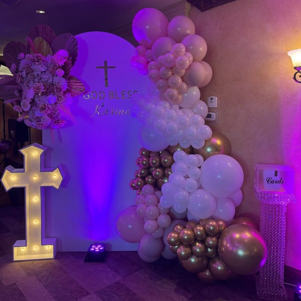 A white backdrop with a gold cross and "God Bless Karina" in gold, adorned with pink and gold balloons.