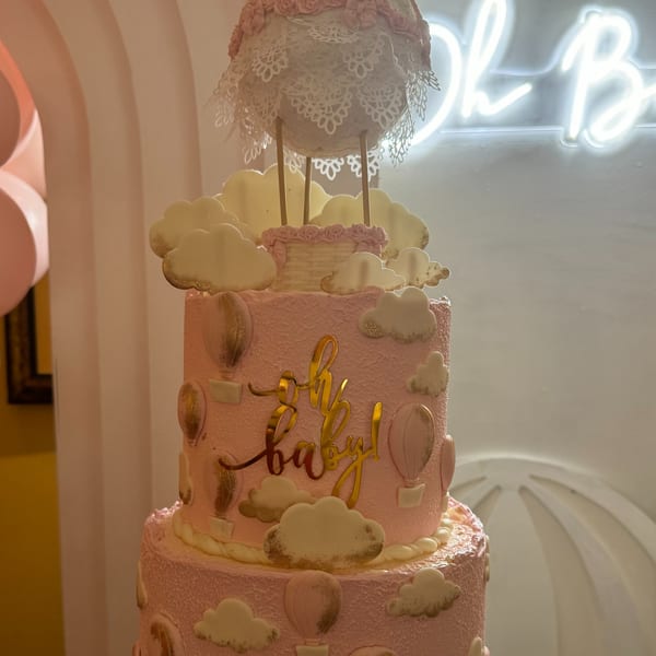 A multi-tiered pink cake with hot air balloons, clouds, and "Oh Baby!" in gold script.