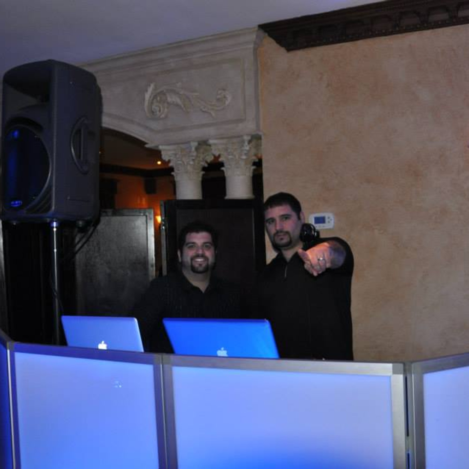 Two DJs with laptops and speakers at Luciano's Ristorante & Lounge, one pointing at the viewer.