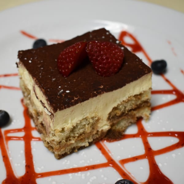 A slice of tiramisu with cocoa powder, two strawberries, and blueberries on a white plate with red sauce.
