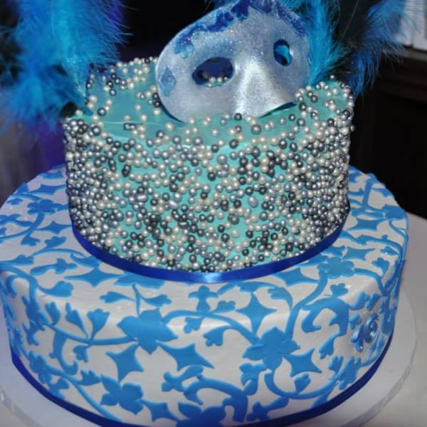 Two-tier cake with blue and white floral patterns, topped with a masquerade mask and feathers.