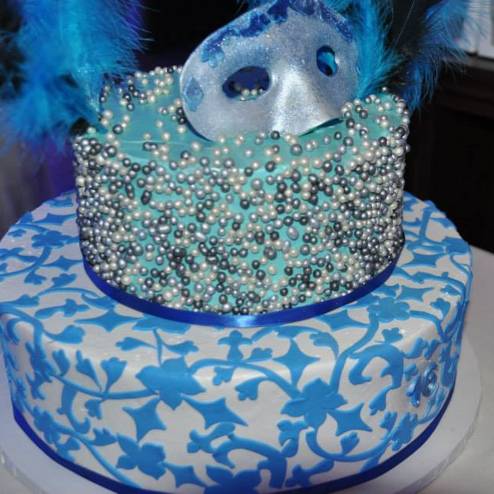 Two-tier cake with blue and white floral patterns, topped with a masquerade mask and feathers.