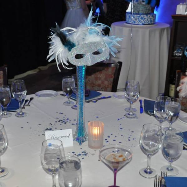 A formal dining table set with glasses, a "Reserved" sign, and a blue feathered masquerade mask centerpiece.