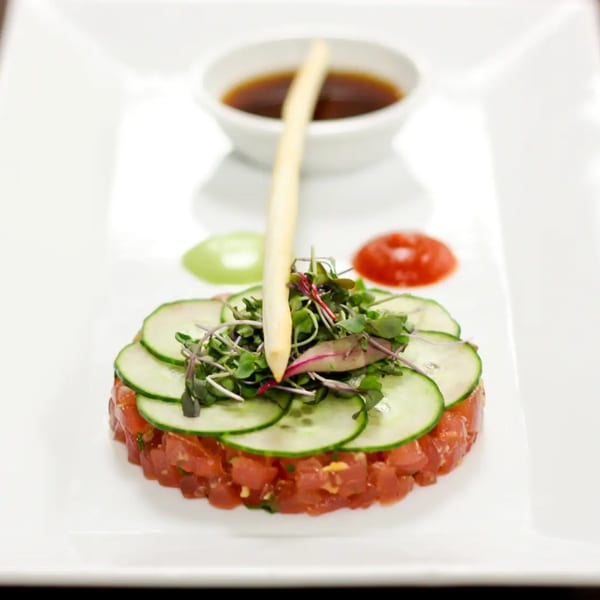 Tuna tartare with cucumber slices, microgreens, a breadstick, and two sauces on a white plate.