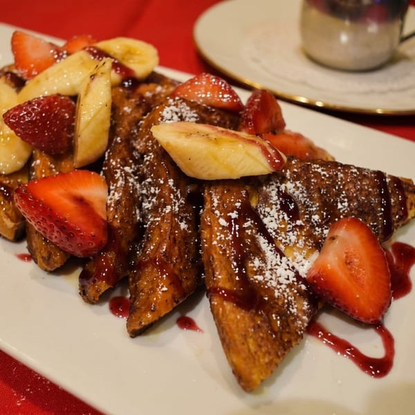Golden French toast topped with sliced strawberries, bananas, powdered sugar, and berry syrup.
