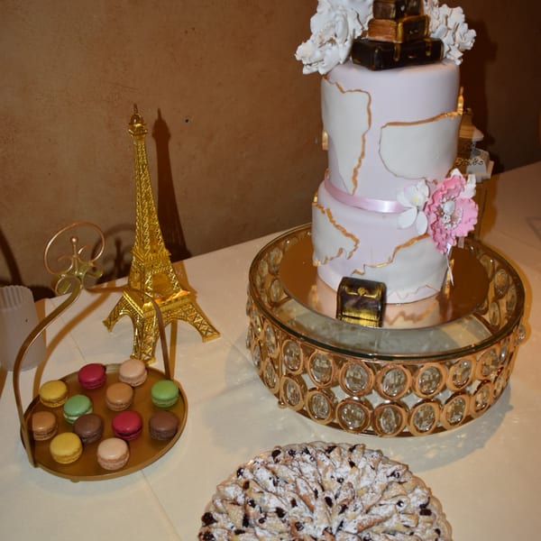 A two-tier cake with gold accents and flowers, macarons, and pastries on a white tablecloth.