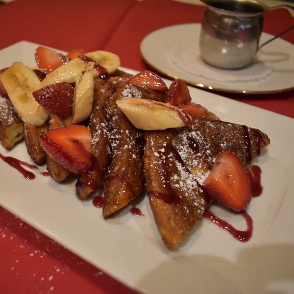 French toast with strawberries, bananas, powdered sugar, and raspberry drizzle on a white plate.