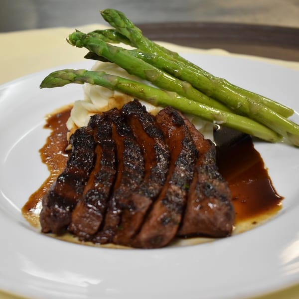 Sliced steak with a rich sauce, mashed potatoes, and green asparagus spears on a white plate.