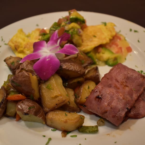 A breakfast plate with scrambled eggs, roasted potatoes, sliced ham, and a purple orchid garnish.