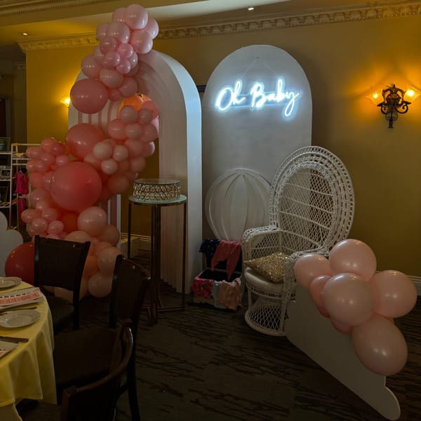 Baby shower decor with pink balloons, a white wicker chair, and a neon "Oh Baby" sign.