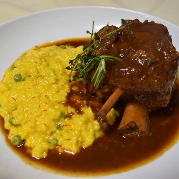 Braised lamb shank with saffron risotto, green peas, and fresh herbs on a white plate.