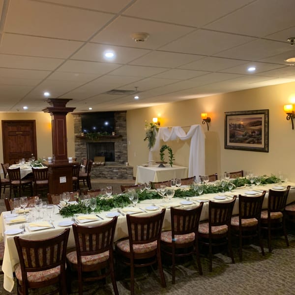 Long dining tables set with white tablecloths, place settings, and green garlands in a warm-lit room.