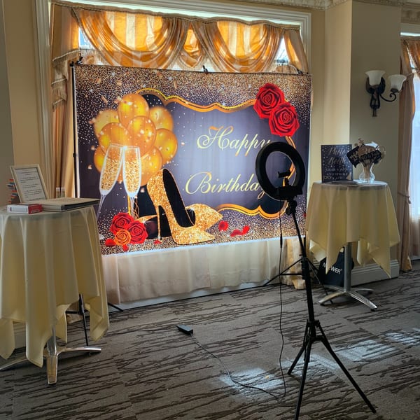 A "Happy Birthday" photo booth backdrop with golden balloons, champagne glasses, and red roses.